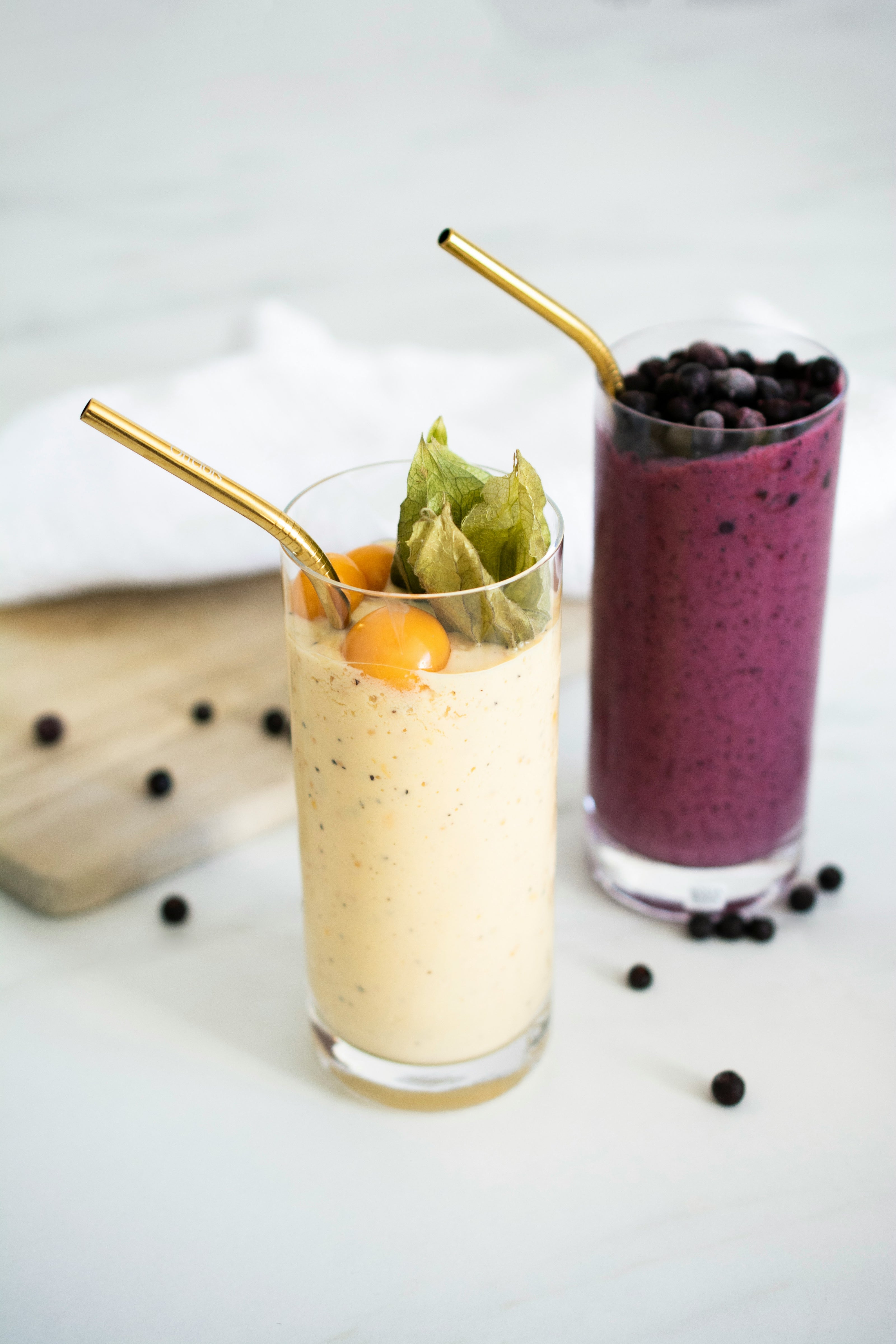 Two smoothies in glass cups with gold straws on a white surface.